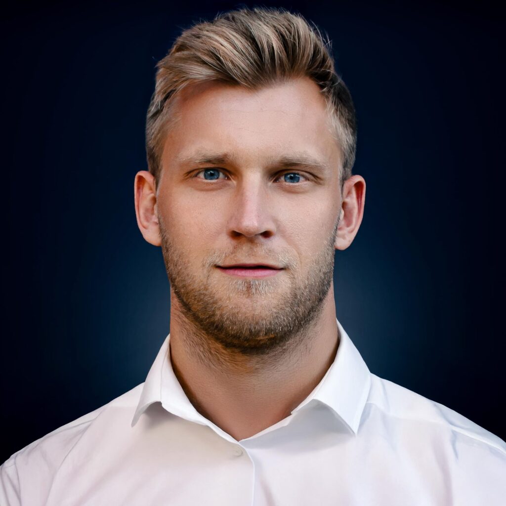Ein Mann mit blonden Haaren, Bart, neutralem Ausdruck, weißem Hemd.
